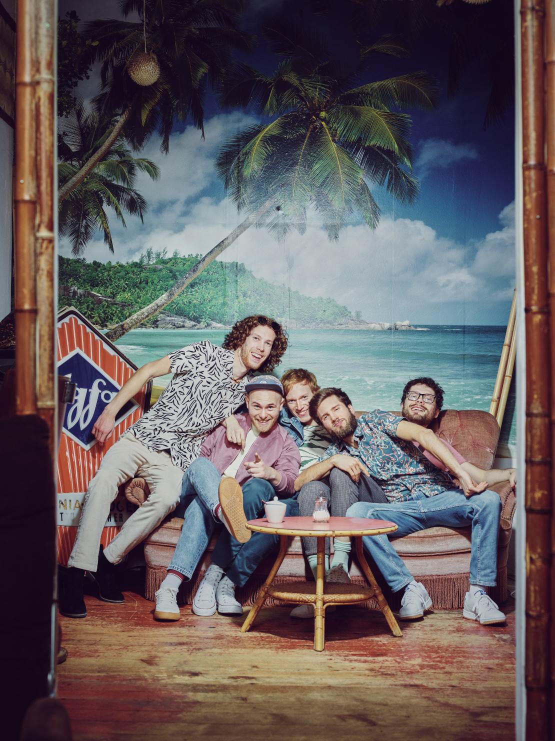 Fünf junge Männder sitzen vor einem Hintergrund mit Strand, blauem Meer und Palmen