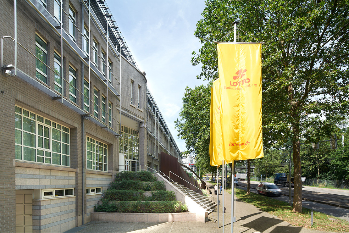 Vor dem Gebäude der Zentrale Lotto Baden-Württemberg mit gelben Lotto-Fahnenmast.