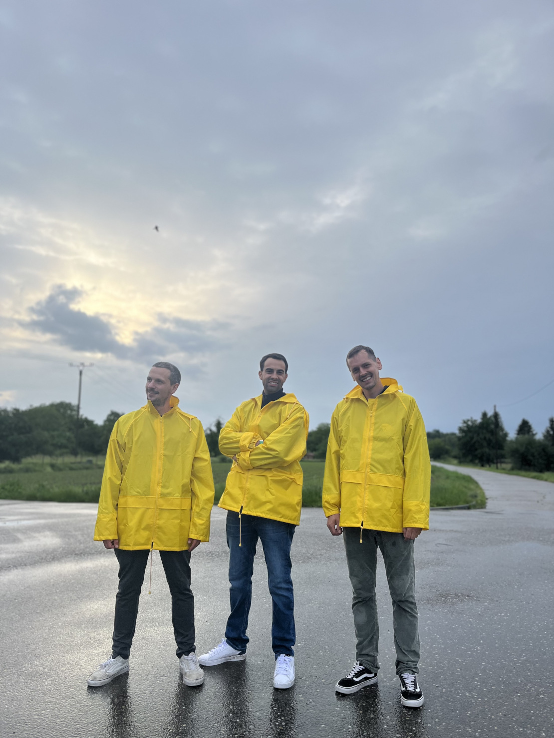 Drei Männer in gelben Regenjacken stehen auf einer nassen Straße mit bewölktem Himmel