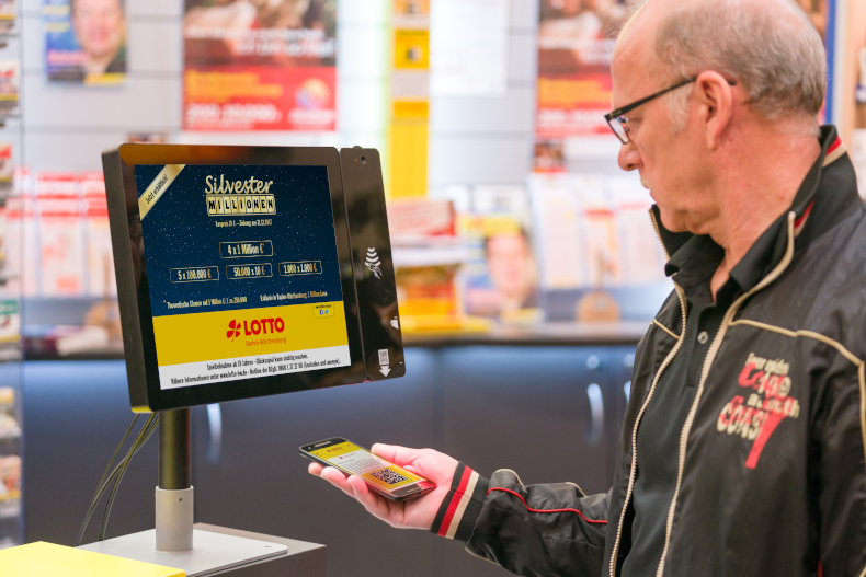 Mann gibt in einer Lotto-Annahmestelle Spielauftrag für Silvestermillionen ab. Er hält den QR-Code der ServiceCard digital unter den Scanner. 